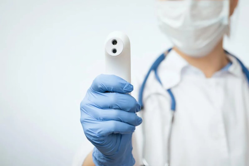 Close-up shot of doctor wearing protective surgical mask ready to use infrared forehead thermometer (thermometer gun) to check body temperature for virus symptoms