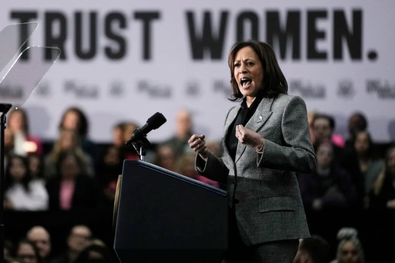 Reproductive Freedom - Harris on stage speaking to a microphone on podium, in front of crowd with banner that reads "trust Women".