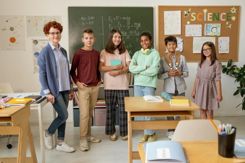 Group of young children along with a teacher standing in front of a chalkboard.
