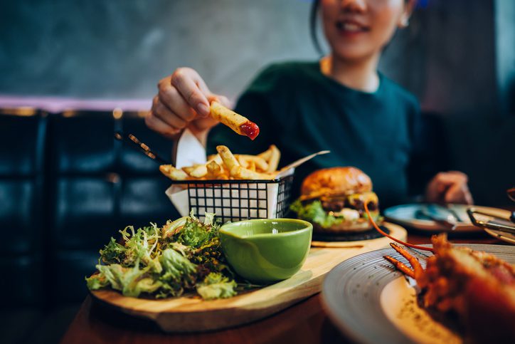 Cropped shot of young Asian woman eating freshly made delicious burger, dipping fries in ketchup on the dining table. Enjoying a feast in the restaurant. People, food and lifestyle